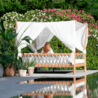 Royal Botania tuskany lounger with teak frame and white fabric, positioned on stone patio near water with floral greenery.