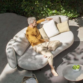 Seated person with gray hair wearing yellow clothing rests on a curved cushioned seat near a small white table with spherical objects