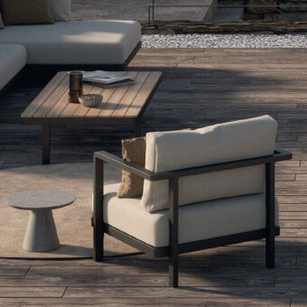 Alura lounge chair, dark metal frame, cream cushions, with wood coffee table, tan pillow, and gray stool on textured rug, outdoor patio scene.