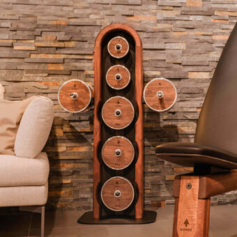 NOHRD weightplate tower with dark wood-grain plates against brown stone veneer, featuring 5.0 kg and 2.5 kg markings, with beige armchair nearby.