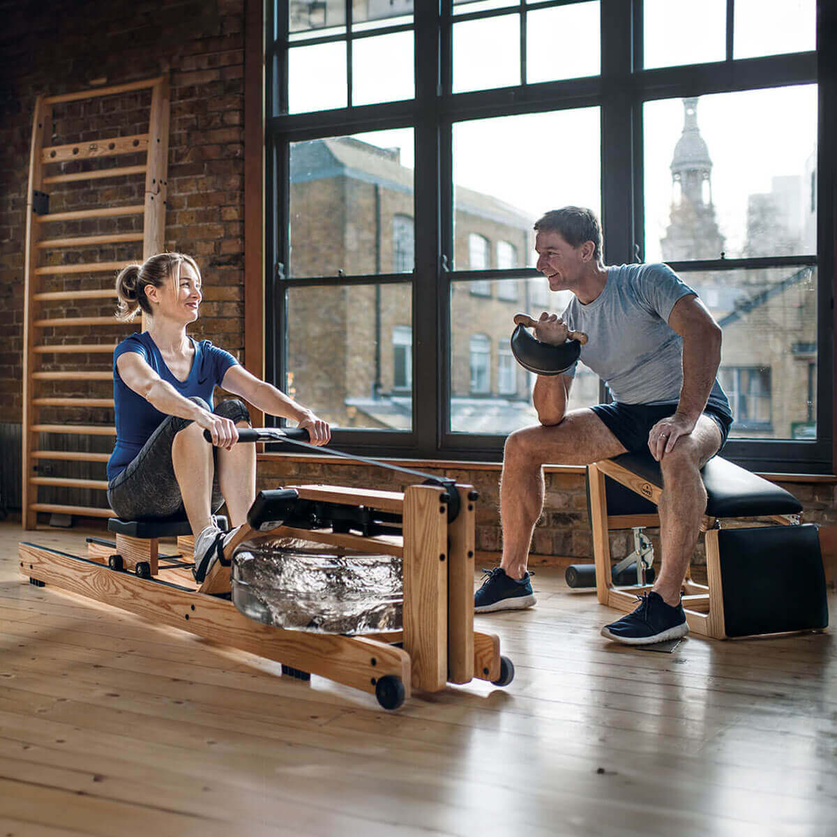 WaterRower Nohr rowing machine with light wood frame and clear tank, alongside a man with dumbbells in a minimalist modern interior.